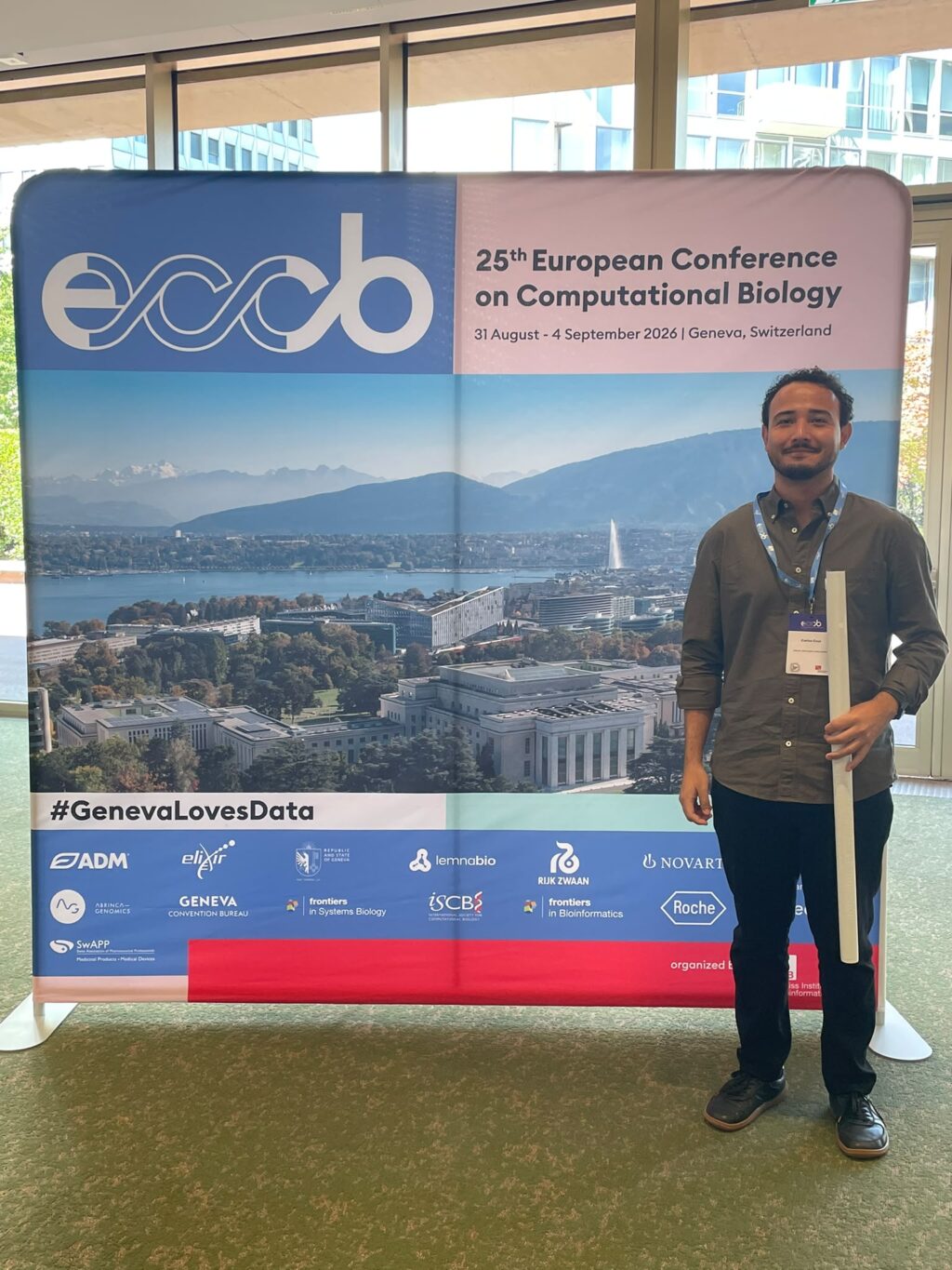 Carlos Cruz junto al banner de su investigación en el Conferencia Europea de Biología Computacional en Ginebra, Suiza.